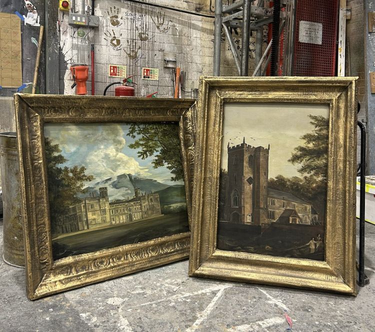 2 large guilt frame regency style landscape paintings of houses are propped up in a paint workshop
